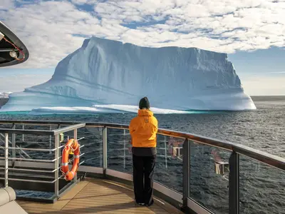 Eine Person in orangefarbener Jacke blickt von Deck der Seabourn Venture auf einen riesigen, blau schimmernden Eisberg im Meer.