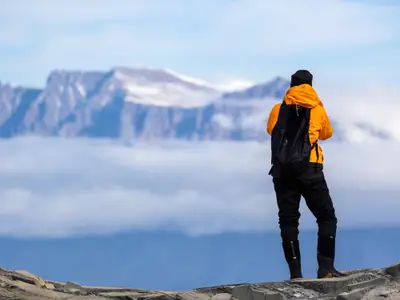 Ein Wanderer in gelber Jacke blickt bei einer Expedition der Seabourn Venture auf schneebedeckte Berge über den Wolken.