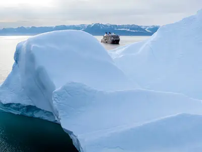 Luftaufnahme der Seabourn Venture im Polargebiet hinter einem gewaltigen weißen Eisberg vor einer schneebedeckten Bergkulisse.