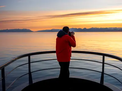 Ein Passagier in einer roten Jacke steht an der Reling an Bord der Seabourn Venture und fotografiert die eisige Bergkulisse bei Sonnenuntergang.