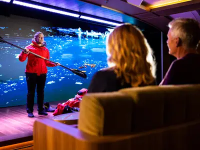 Gäste verfolgen an Bord der Seabourn Venture einen Vortrag über Kajaktouren vor einer großen Videowand mit arktischer Landschaft.