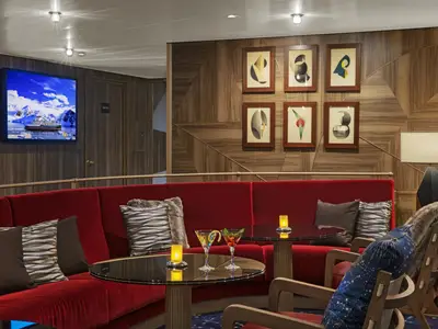 Eine gemütliche Lounge an Bord der Seabourn Venture mit roten Polstersesseln, Glastischen, Cocktails und moderner Kunst an den Holzwänden.