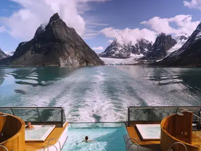 Blick vom Heck der Seabourn Venture mit Infinity-Pool und Whirlpools auf das Kielwasser und schneebedeckte Berge in Grönland.