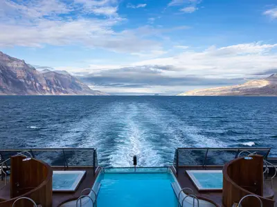Blick über den Infinity-Pool und zwei Whirlpools am Heck an Bord der Seabourn Venture auf ein tiefblaues Meer und hohe Berge.