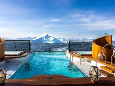 Infinity-Pool und Whirlpools an Bord der Seabourn Venture mit Blick auf einen riesigen Eisberg und schneebedeckte Berge in kalten Gewässern.