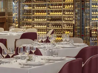 Eingedeckte Tische mit Weingläsern vor einem großen Weinschrank an Bord der Seabourn Venture.