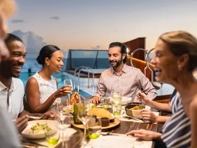 Eine fröhliche Gruppe genießt ein luxuriöses Abendessen unter freiem Himmel an Bord der Seabourn Venture bei Sonnenuntergang.