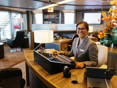 Eine lächelnde Mitarbeiterin mit Brille und Namensschild am modernen Empfangstisch an Bord der Seabourn Venture.
