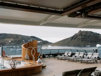 Das luxuriöse Sonnendeck an Bord der Seabourn Venture mit Whirlpool und Liegestühlen bietet Ausblick auf Eisberge und raue Berge in der Antarktis.