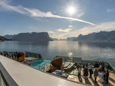 Gäste entspannen am Pool und den Whirlpools auf dem Sonnendeck der Seabourn Venture inmitten einer grönländischen Fjordlandschaft bei Sonnenschein.