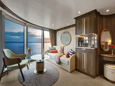 Wohnbereich einer Suite an Bord der Seabourn Venture mit Sofa, Sessel, Tisch und großem Fenster mit Meerblick bei Sonnenuntergang.