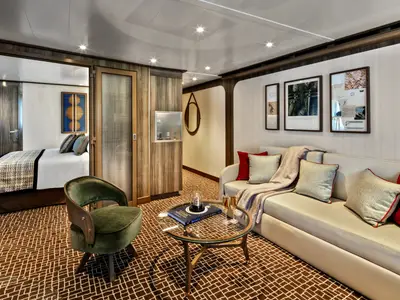 Wohnbereich einer Suite an Bord der Seabourn Venture mit hellem Sofa, grünem Sessel, Couchtisch aus Glas und Blick ins Schlafzimmer.