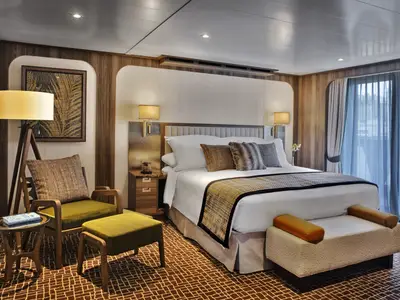 Blick in eine elegant gestaltete Suite an Bord der Seabourn Venture mit großem Bett, Holzpaneelen, Sesseln und Balkontür.