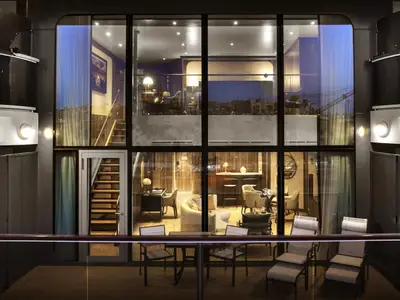 Blick von außen durch die große Fensterfront in eine luxuriöse, beleuchtete zweistöckige Suite an Bord der Seabourn Venture.