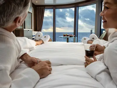 Ein Paar in weißen Bademänteln entspannt auf dem Bett in einer Suite an Bord der Seabourn Venture und blickt aufs Meer.