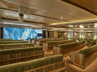 Moderner Vortragssaal an Bord der Seabourn Venture mit geschwungenen, grünen Polsterbänken vor einer großen Leinwand mit Eisberg-Motiv.