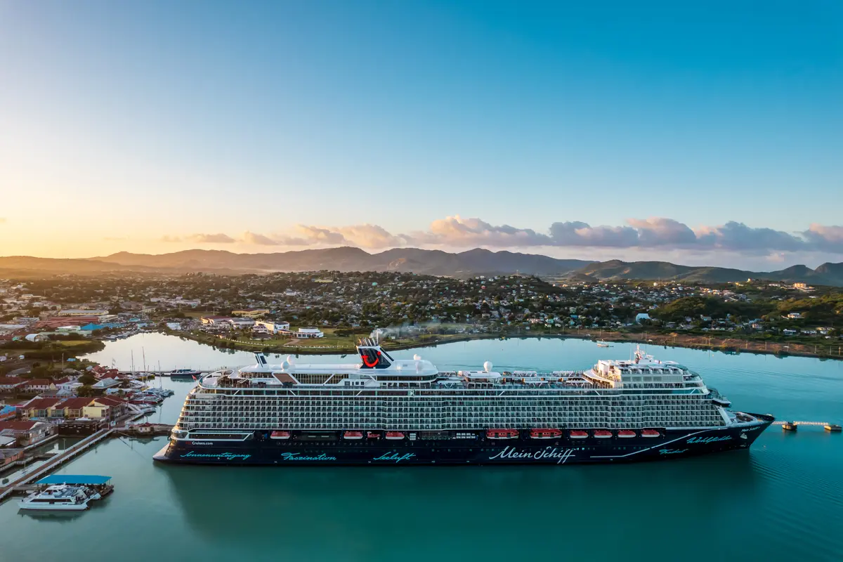 Die Mein Schiff 1 liegt in einem türkisfarbenen Hafen vor einer Küstenstadt mit grünen Bergen im Hintergrund bei goldenem Sonnenuntergang. Ein atemberaubendes Panorama.