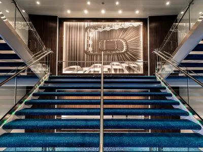Zentrale Treppe an Bord der Mein Schiff 1 mit blauem Teppich, Glasgeländern und einem großen, beleuchteten Kunstwerk an der Wand, das eine Wendeltreppe von oben zeigt. Grüne LED-Lichter säumen die Stufen.