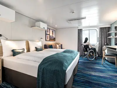 Barrierefreie Aussenkabine an Bord der Mein Schiff 1. Eine Frau im Rollstuhl blickt aus dem Fenster aufs Meer. Die Kabine hat ein Doppelbett, Sitzecke und Schreibtisch.