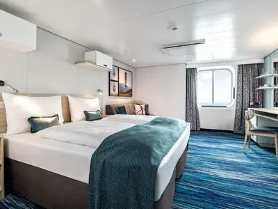 Detailansicht einer modernen, barrierefreien Außenkabine an Bord der Mein Schiff 1 mit hellen Möbeln, blauen Teppich und einem großen Fenster zum Meer.