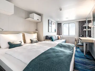 Geräumige und moderne Außenkabine an Bord der Mein Schiff 1. Zwei Einzelbetten, eine Couch und ein Schreibtisch sind zu sehen. Ein großes, rundes Fenster bietet Meerblick.