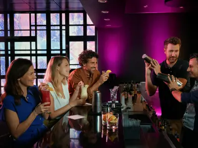Vier Freunde bei einem Cocktail-Workshop in einer stilvollen Bar an Bord der Mein Schiff 1. Zwei lernen vom Barkeeper, die anderen genießen Drinks. Warme magenta-Beleuchtung schafft eine lebhafte Atmosphäre.