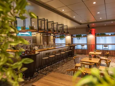 Blick in die rustikale Ebbe & Flut Bier Bar an Bord der Mein Schiff 1. Eine lange Holztheke mit Barhockern und Zapfanlagen prägt den Raum. Rechts davon stehen Holztische mit Stühlen. Viele Flaschen und beleuchtete Fenster sind sichtbar.