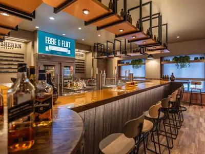 Die Ebbe & Flut Bier Bar an Bord der Mein Schiff 1. Eine moderne Bar mit glänzender Theke, Zapfanlagen, Flaschen und Barhockern. Eine Leuchtschrift zeigt den Namen.