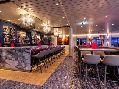 Die elegant eingerichtete LUMAS Bar an Bord der Mein Schiff 1, mit einem langen Marmortresen und gepolsterten Barhockern. Im Hintergrund sind Spirituosenregale und Kronleuchter zu sehen. Die Atmosphäre ist gedämpft und modern.