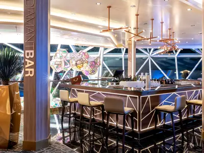 Blick in die stilvolle Diamant Bar an Bord der Mein Schiff 1 mit beleuchteter Theke und bequemen Barhockern. Im Hintergrund schimmern farbige, geometrische Leuchten. Die Bar ist leer.
