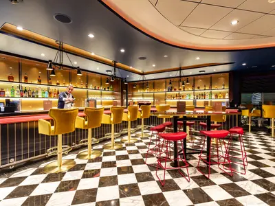 Die moderne Schau Bar an Bord der Mein Schiff 1 mit einem Barkeeper, der einen Cocktail zubereitet. Die Bar hat goldene Regale mit Spirituosen, gelbe Samthocker und einen schwarz-weiss karierten Boden.