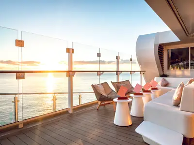 Blick auf die Hoheluft Bar an Bord der Mein Schiff 1 bei Sonnenaufgang. Moderne Sitzmöbel und Barhocker mit Blick auf das Meer und den Horizont.