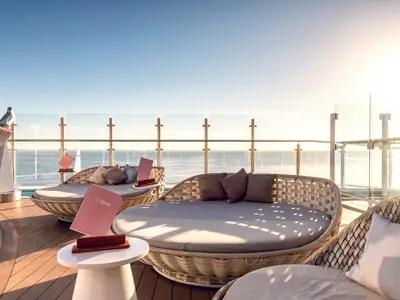 Die Hoheluft Bar an Bord der Mein Schiff 1 mit gemütlichen Lounge-Betten und rosa Menükarten. Im Hintergrund glitzert das Meer unter strahlendem Sonnenschein.