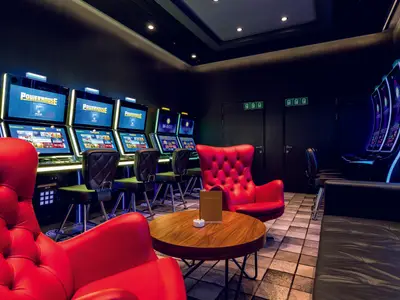 Eine moderne Casino-Lounge an Bord der Mein Schiff 1. Mehrere Reihen von hell beleuchteten Spielautomaten stehen bereit, ergänzt durch bequeme rote Sessel und eine schwarze Ledercouch. Ein kleiner Holztisch mit Ascher und Broschüre steht in der Mitte.