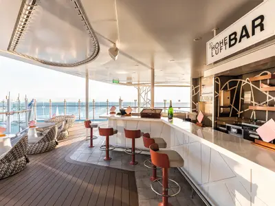 Blick auf die moderne Hoheluft Bar an Bord der Mein Schiff 1 im sanften Licht des Sonnenuntergangs. Die Bar mit weissen Theken und roten Hockern steht auf einem Deck mit Meerblick.