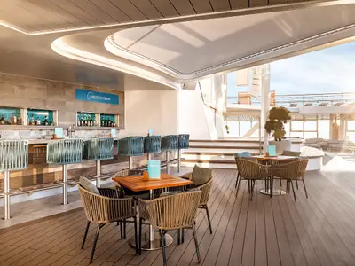 Die moderne Unverzicht Bar an Bord der Mein Schiff 1. Sie bietet Hocker an der Bar und Tische mit Rattanstühlen auf einem großzügigen Holzdeck. Hell beleuchtet vom Sonnenlicht.