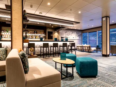 Gemütliche "Ruhepol" Bar-Lounge an Bord der Mein Schiff 1. Sie zeigt eine lange Bar, Sitzgruppen mit Sofas und einen Ausblick auf grüne Hügel durch große Fenster.