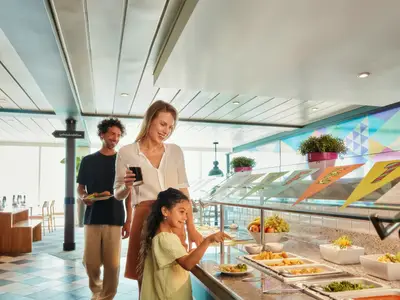 Eine lächelnde Familie mit Mutter, Vater und Kind wählt Speisen am Buffet an Bord der Mein Schiff 1. Reichhaltige Auswahl an frischen Früchten und warmen Gerichten ist zu sehen.