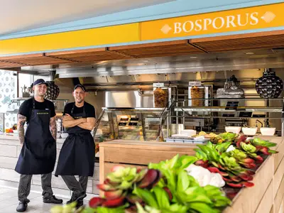 Zwei laechelnde Crewmitglieder der Mein Schiff 1 in Schuerzen vor der Bosporus Snackbar. Doener-Spiessse und frische Zutaten im Buffetbereich sichtbar.