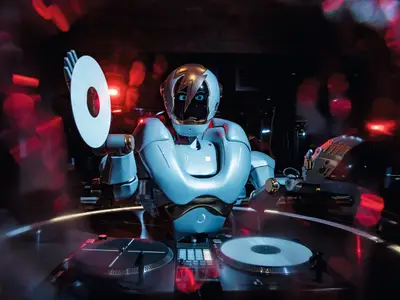 Ein Roboter-DJ mit blauen Augen und Blitzsymbol auf dem Helm legt Platten auf zwei Turntables an Bord der Mein Schiff 1 auf. Rote Lichter erzeugen eine Club-Atmosphäre.