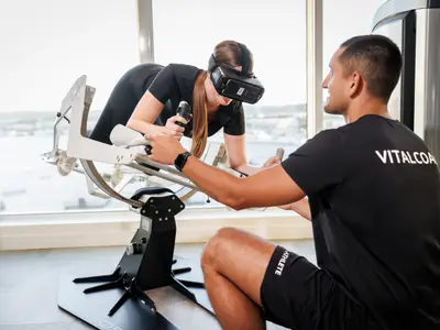 Eine Frau mit VR-Brille trainiert an einem speziellen Fitnessgerät unter Anleitung eines Trainers an Bord der Mein Schiff 1.