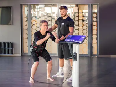 An Bord der Mein Schiff 1 macht ein Mann in EMS-Kleidung Kniebeugen unter Anleitung eines Personal Trainers. Das EMS-Steuergerät steht im hellen Fitnessbereich bereit.