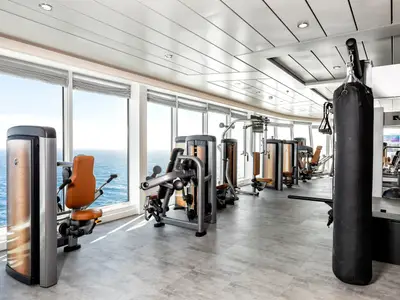 Blick in den hellen Fitnessraum an Bord der Mein Schiff 1 mit zahlreichen modernen Kraftgeräten und einem Boxsack. Grosse Fenster bieten Panoramablick auf das Meer und den Horizont.