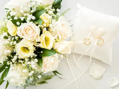 Ein festlicher Hochzeitsstrauß mit weißen und cremefarbenen Rosen liegt neben einem weißen Ringkissen mit goldenen Eheringen, bereit für eine Trauung an Bord der Mein Schiff 1.