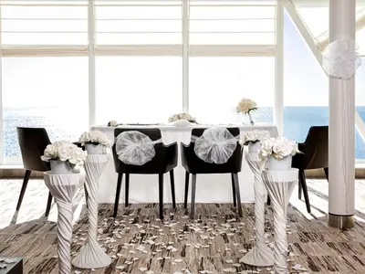 Ein festlich dekorierter Innenbereich an Bord der Mein Schiff 1 für eine Hochzeit. Zwei Stühle mit Schleifen und ein Tisch mit weißen Blumen stehen vor großen Fenstern mit Meerblick. Weiße Blütenblätter liegen auf dem Boden.