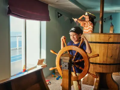 Zwei Kinder in Piratenkostümen spielen begeistert in einem nachgebauten Piratenschiff an Bord der Mein Schiff 1. Ein Junge steht am Steuerrad, ein Mädchen blickt von oben.