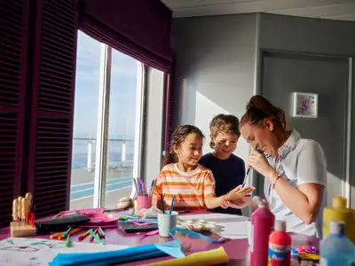 Eine Crew-Mitarbeiterin malt mit zwei Kindern im Kids-Club an Bord der Mein Schiff 1. Sie bemalen die Hand eines Mädchens, während ein Junge zusieht. Im Hintergrund ist das Meer sichtbar.