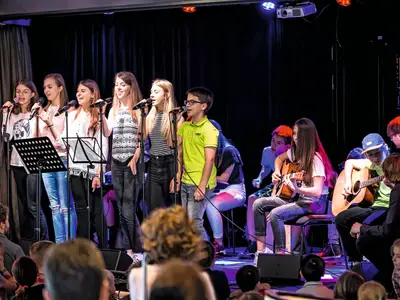 Eine Gruppe von Kindern und Jugendlichen singt und spielt Gitarre auf einer hell beleuchteten Bühne an Bord der Mein Schiff 1 vor einem Publikum.