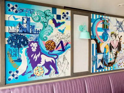 Farbintensive Wandbilder mit fantasievollen Motiven wie Löwe, Schloss und Bäckerin an Bord der Mein Schiff 1. Dazu mintfarbene Leuchte und lila Sitzbank.
