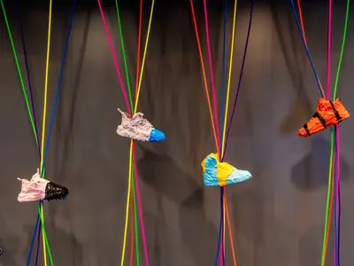 Eine farbenfrohe Kunstinstallation an Bord der Mein Schiff 1 zeigt mehrere, mit Farbe überzogene Turnschuhe, die an bunten Seilen von der Decke hängen. Ein modernes, lebhaftes Designelement.